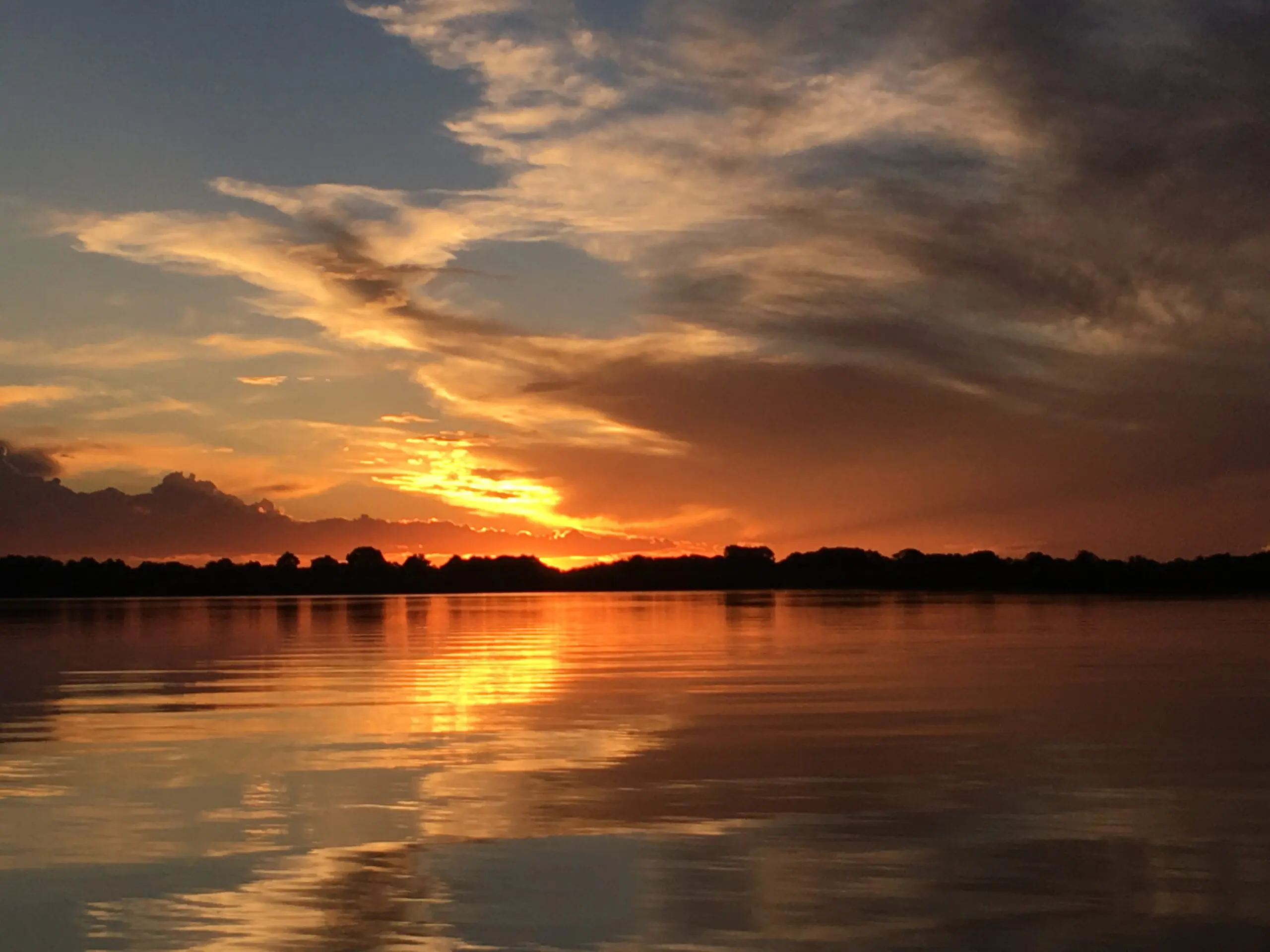 Een levendige zonsondergang boven een kalm meer, met dramatische wolken en oranje, gele en paarse tinten die weerspiegelen op het wateroppervlak. Het donkere silhouet van bomen omlijnt de horizon.
