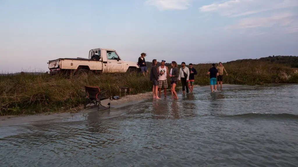 Een groep mensen verzamelt zich aan de rand van een meer of rivier bij een met gras begroeide oever, met een geparkeerde witte pick-up truck en kampeerspullen in de buurt tijdens zonsondergang. Sommige mensen staan in het ondiepe water, anderen op de oever.
