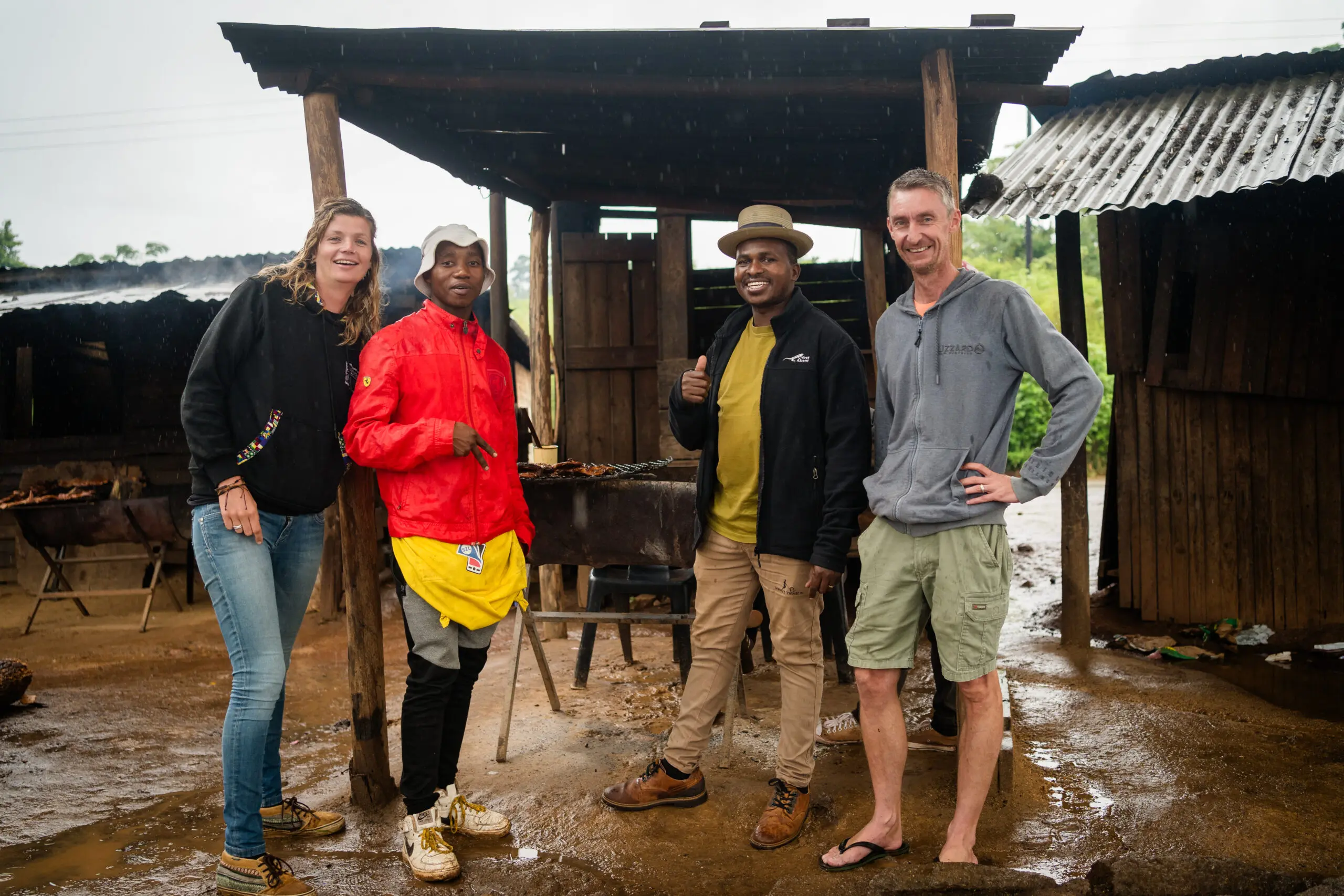 Vier mensen staan en glimlachen samen voor een rustiek houten eetstalletje op een regenachtige dag. De grond is modderig en twee van hen dragen een hoed, terwijl één persoon een korte broek en slippers draagt. Op de achtergrond is gegrild eten te zien.