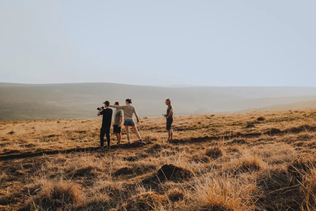 Four people stand on a dry, grassy hill under a pale sky, looking out over a vast, open landscape with rolling hills in the distance.
