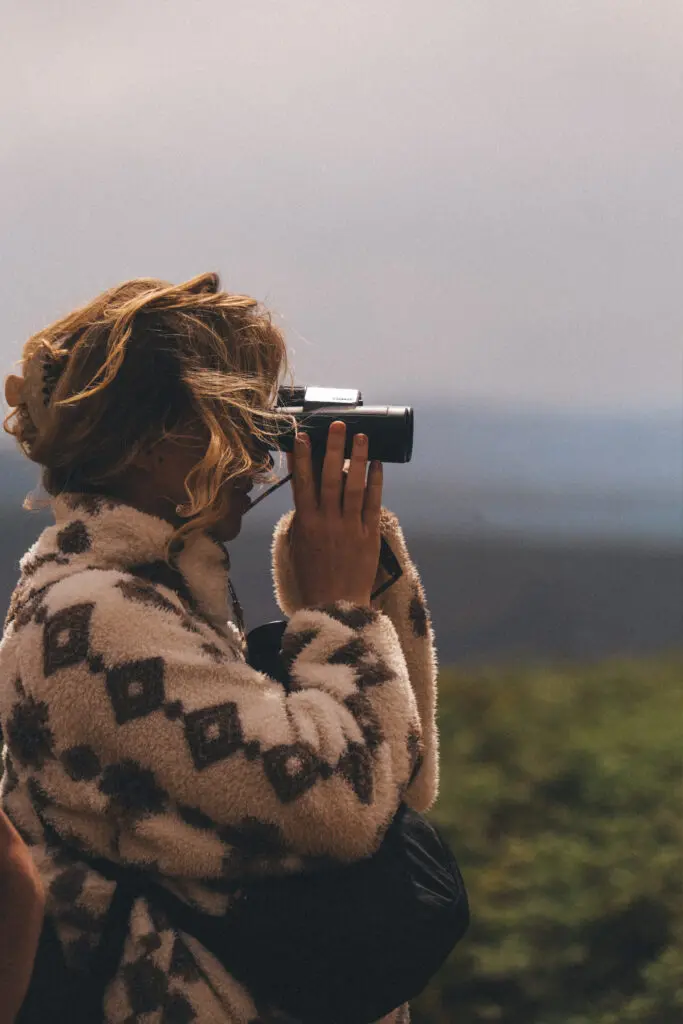 Een persoon met een fleecejack met patroon kijkt door een verrekijker naar buiten, met wazig groen en een bewolkte lucht op de achtergrond.
