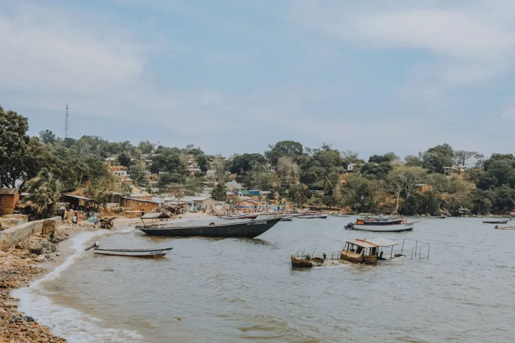Boten liggen aangemeerd langs een kustlijn met verspreide huizen en bomen op de achtergrond; mensen lopen langs het water onder een gedeeltelijk bewolkte hemel.
