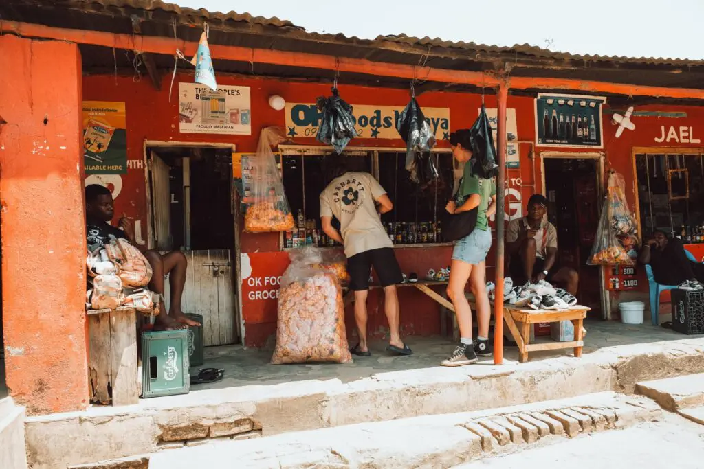 Mensen staan voor een klein, roodgeschilderd winkeltje langs de weg met snacks in zakken en uitgestalde goederen. De winkel heeft open ramen en uithangborden; sommige mensen zitten, terwijl anderen aan het winkelen zijn in een zonnige, buitenomgeving.