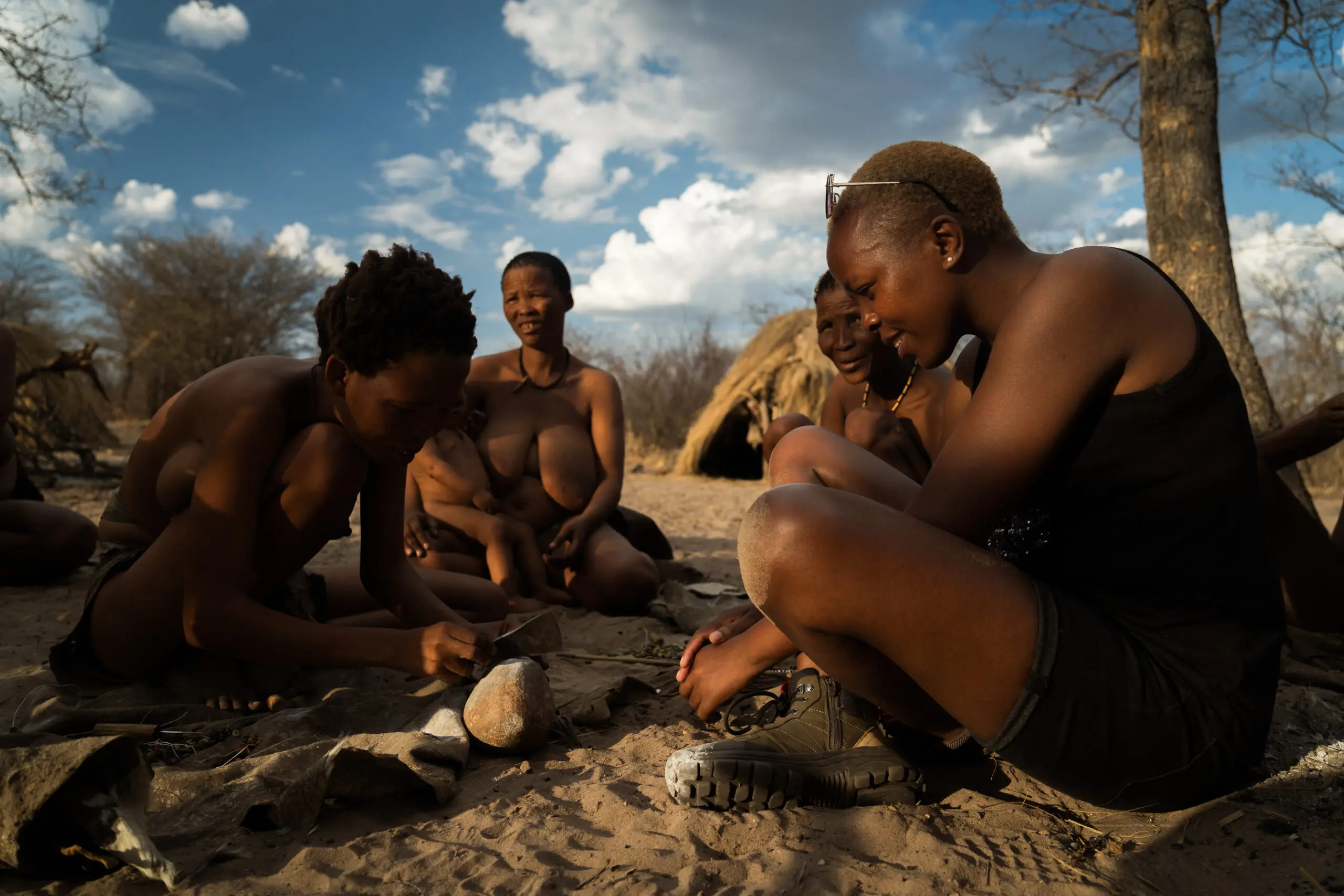 Een groep mensen zit buiten op de grond en één persoon gebruikt gereedschap op een steen. Op de achtergrond zijn hutten en bladerloze bomen te zien onder een gedeeltelijk bewolkte hemel.