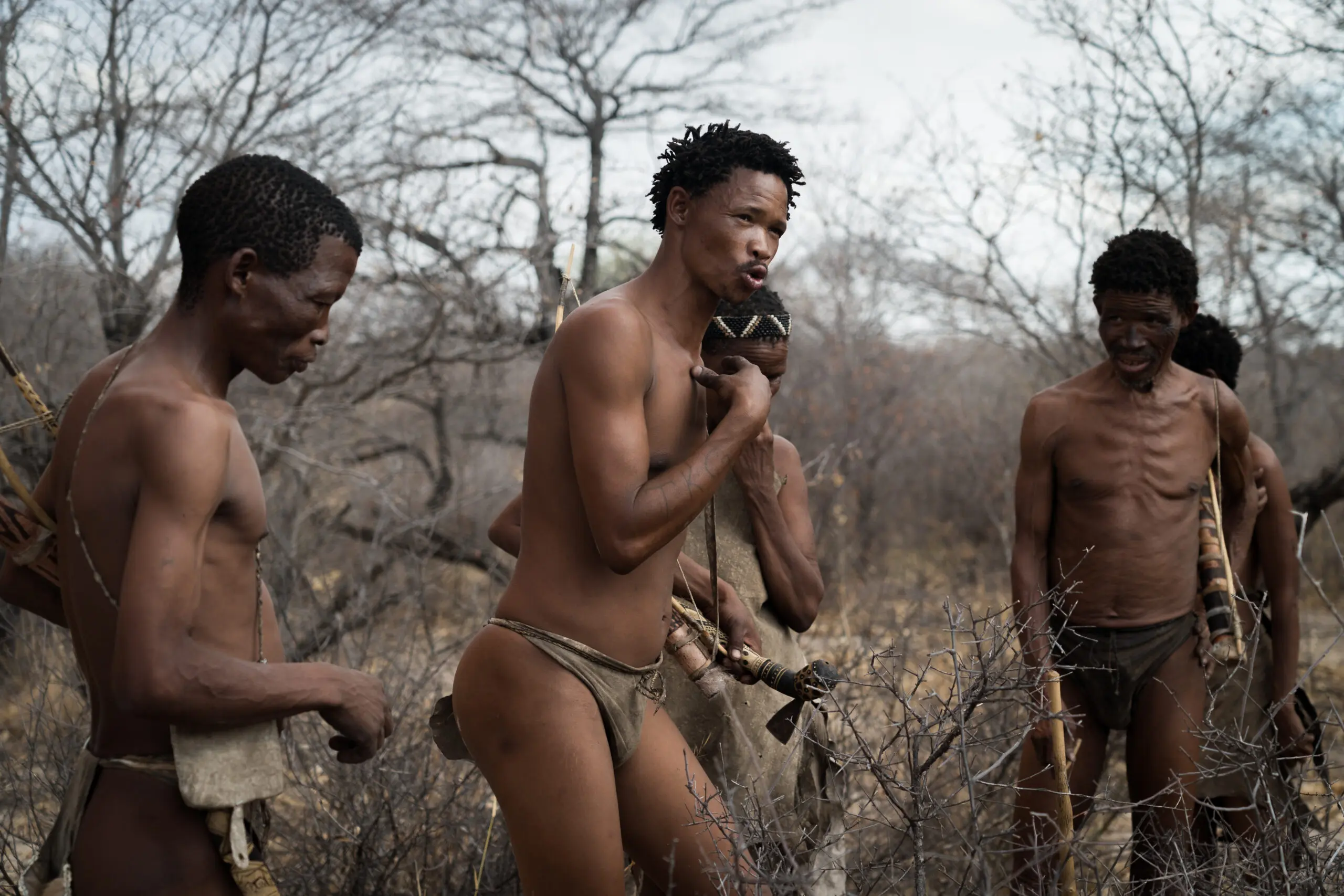 Vier mannen in traditionele lendendoeken staan buiten in een droog, bladerloos landschap en lijken samen te praten of een activiteit te ondernemen. De scène suggereert een culturele of gemeenschappelijke omgeving.