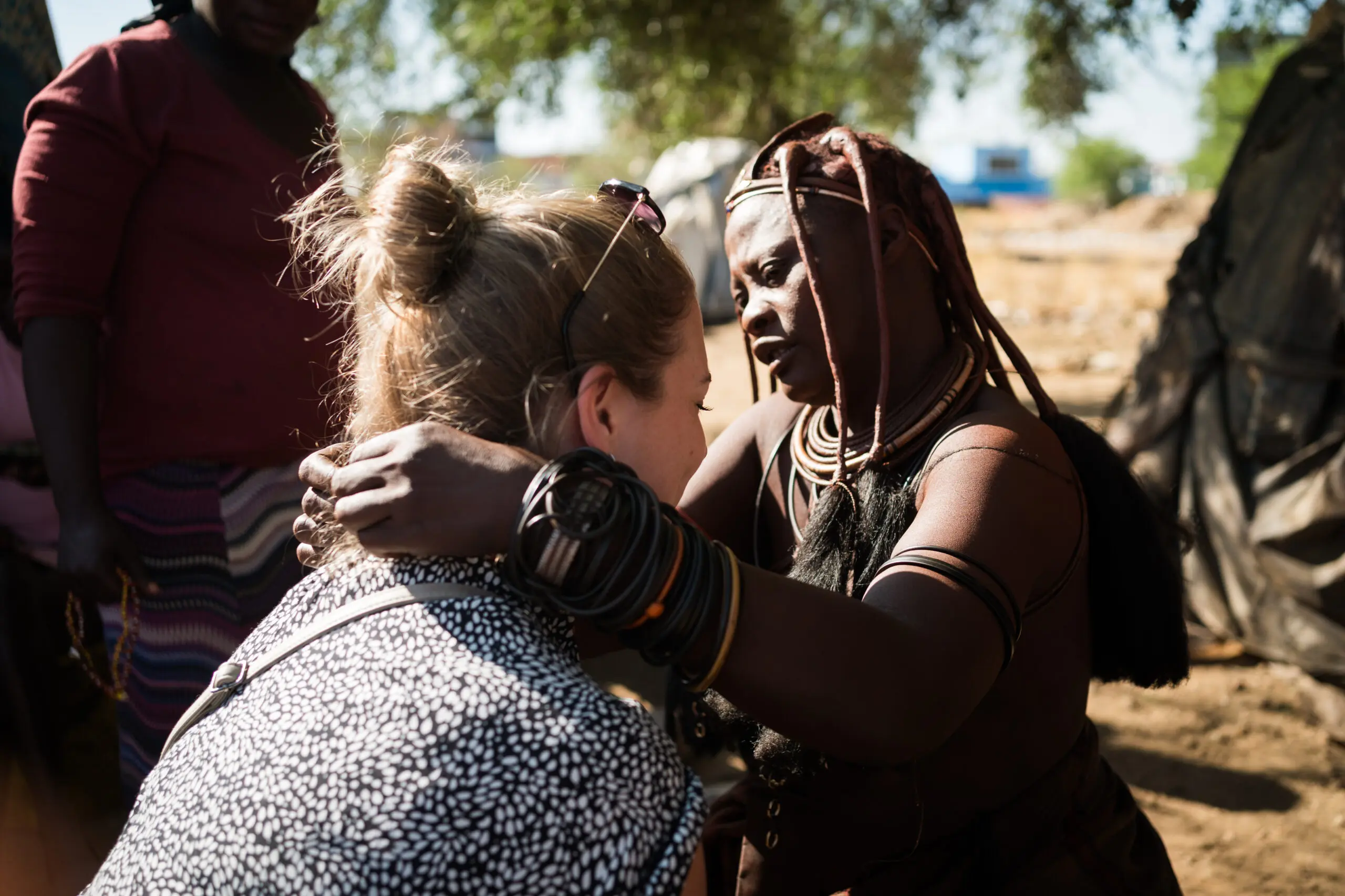 Een Himba-vrouw, getooid met traditionele sieraden en haarversieringen, houdt voorzichtig de schouders van een bezoeker vast terwijl ze in haar ogen kijkt, terwijl een andere persoon op de achtergrond in de buurt staat.
