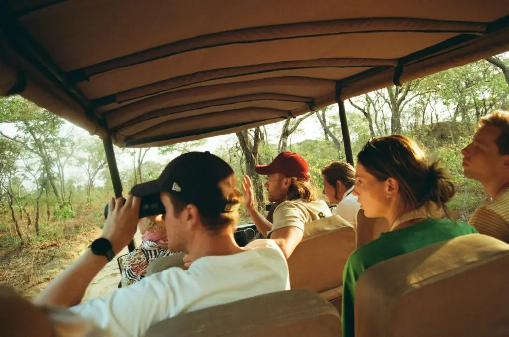 Een groep mensen zit in een safarivoertuig dat door een bebost gebied rijdt. Eén persoon gebruikt een verrekijker, terwijl anderen vooruit kijken, sommigen met hoeden en zonnebrillen op. Zonlicht filtert door de bomen.