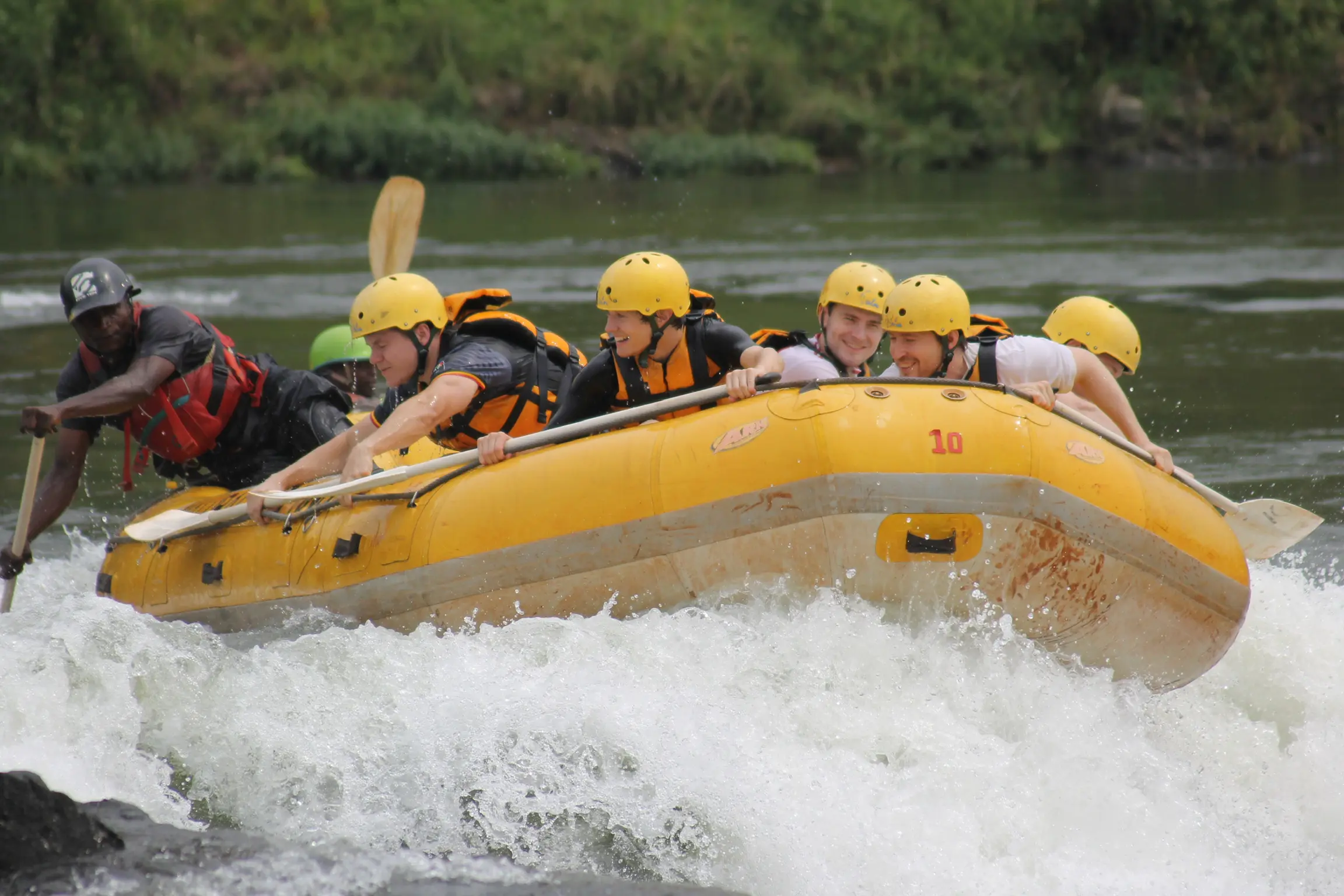Zes mensen met gele helmen en reddingsvesten varen op een geel opblaasbaar vlot door woelig water met een gids voorop. Groene vegetatie omzoomt de rivieroever op de achtergrond.
