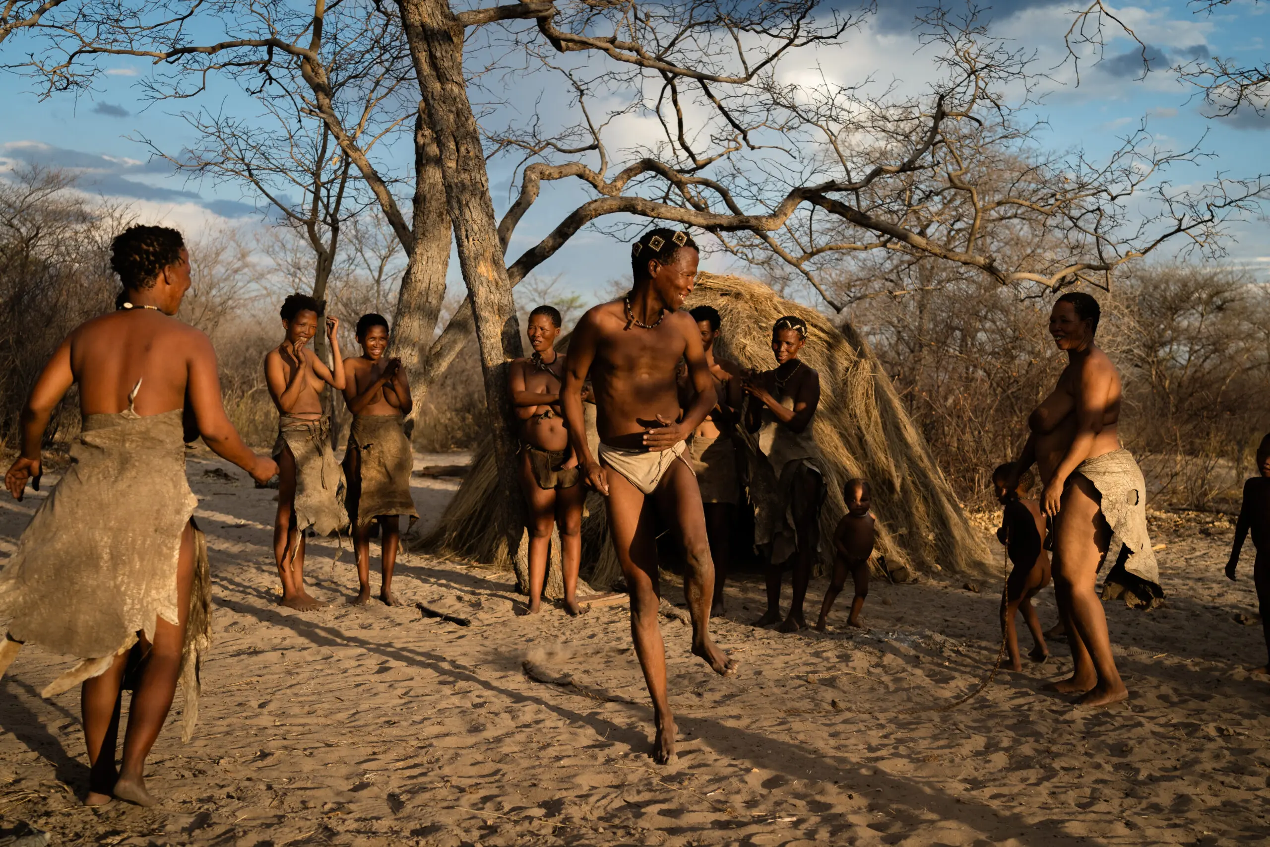 Een groep mensen, gekleed in traditionele kledij, is buiten bijeen. Sommigen klappen en glimlachen, terwijl één persoon in het midden danst. Kale bomen en een rieten afdak zijn zichtbaar op de achtergrond onder een gedeeltelijk bewolkte hemel.