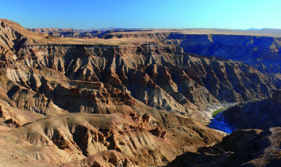 Een uitgestrekte, ruige canyon met steile, gelaagde rotswanden en een kronkelende rivier op de bodem, onder een helderblauwe hemel. Het landschap is dor en dramatisch, met diepe kloven en zonovergoten kliffen.