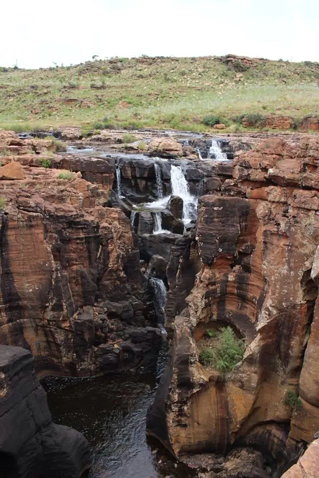 Een smalle waterval cascadeert tussen hoge, gelaagde rode en bruine rotsformaties en eindigt in een kleine donkere poel. Bovenop en binnenin de rotsachtige kliffen groeit schaarse groene vegetatie, met op de achtergrond grasheuvels.