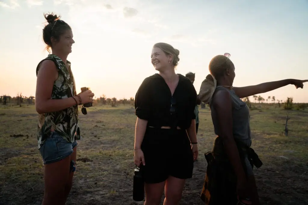 Drie vrouwen staan buiten bij zonsondergang, lachend en pratend. De ene houdt een verrekijker vast, de andere een waterfles en de derde wijst in de verte. De scène is helder met een graslandschap op de achtergrond.