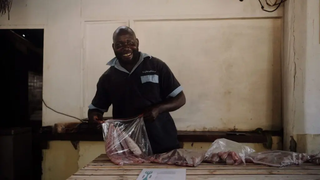 Een lachende man in een donker shirt staat achter een houten tafel en behandelt stukken vlees in een doorzichtige plastic zak. Op de tafel liggen verschillende zakken vlees en de achtergrond toont een effen, lichtgekleurde muur.