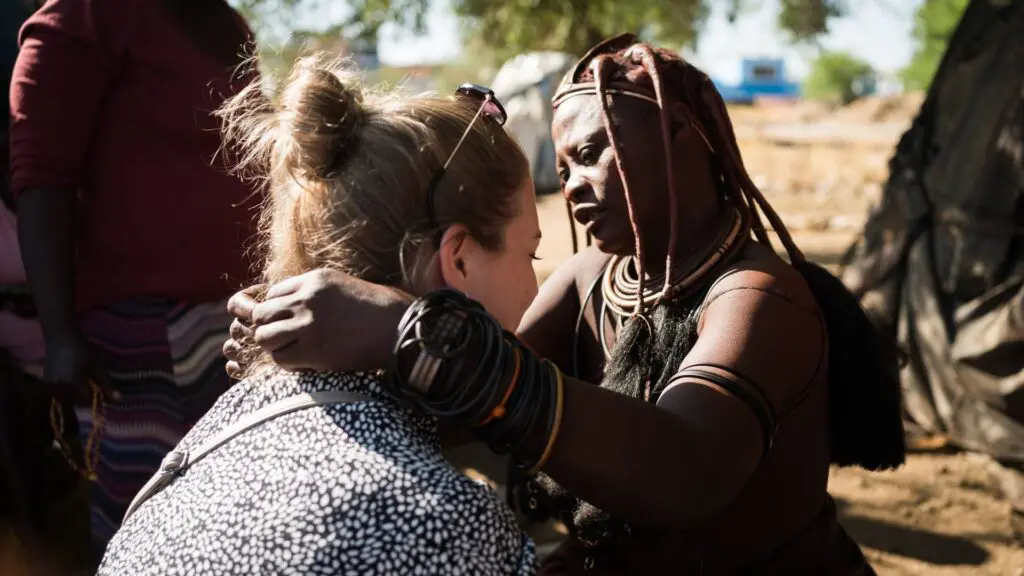 Een Himba vrouw met traditionele juwelen en kleding raakt zachtjes de schouders aan van een zittende vrouw met een lichte huid en blond haar, die van de camera weg kijkt, in een buitenopstelling.