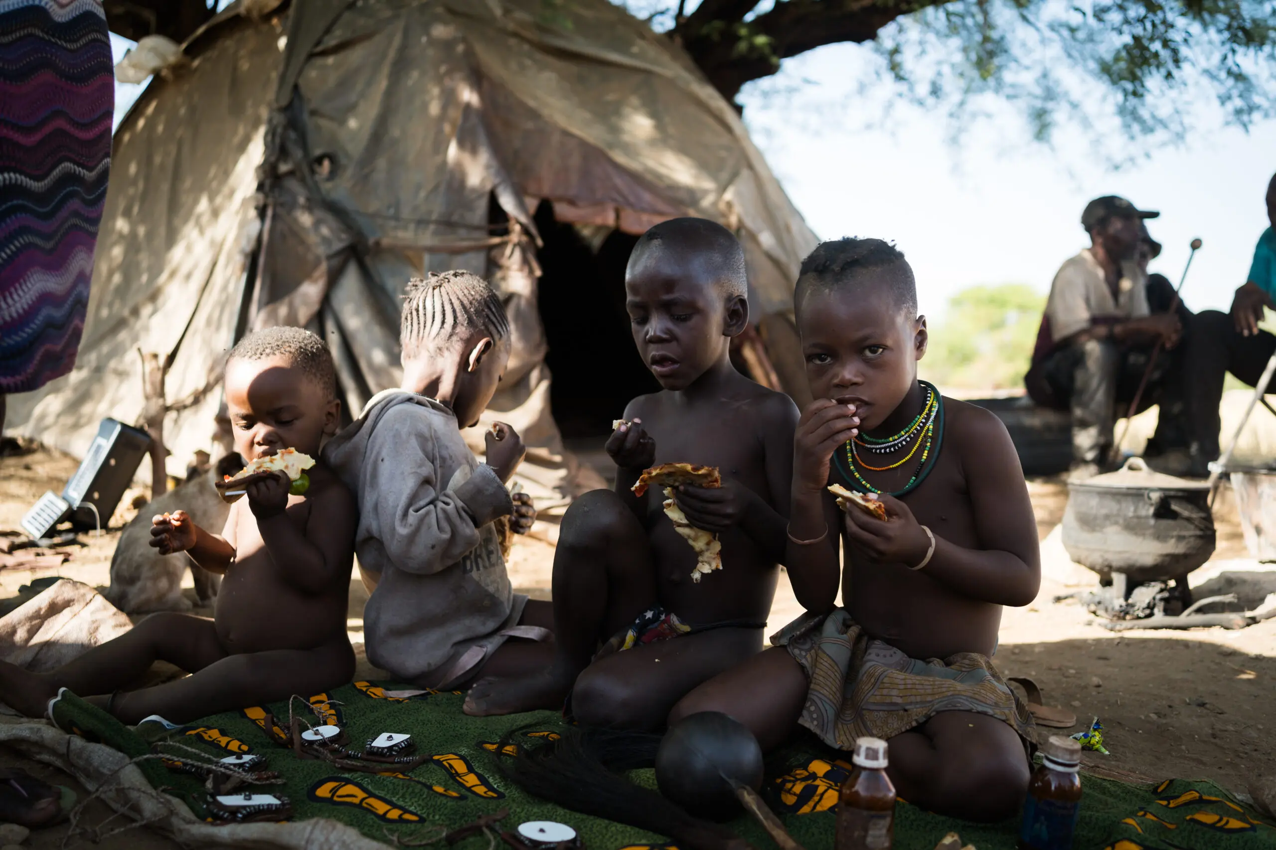 Vier jonge kinderen zitten buiten op een matje bij een tent te eten. Twee volwassenen zijn op de achtergrond te zien bij een pot boven een vuur. Het tafereel wordt overschaduwd door een grote boom.