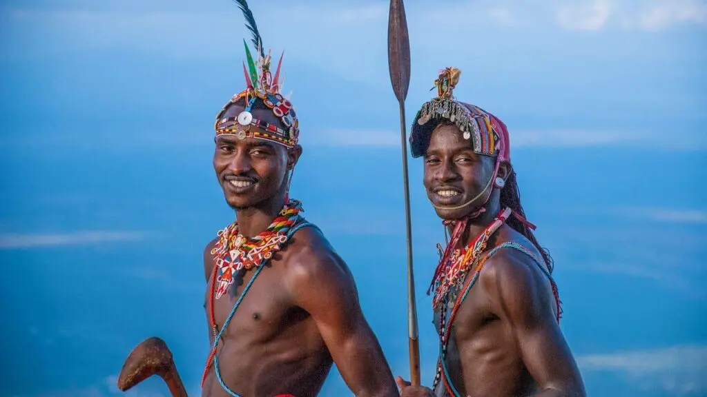 Twee Maasai mannen staan naast elkaar en glimlachen, dragen traditionele kralen sieraden en hoofddeksels, de een houdt een speer vast en de ander een houten knots, met een uitgestrekte blauwe lucht op de achtergrond.