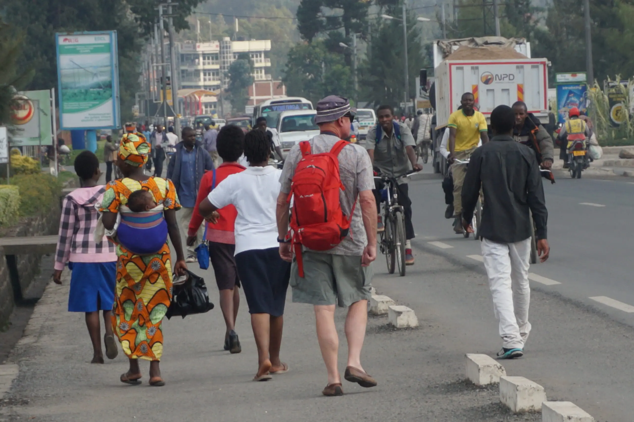 Mensen lopen langs een drukke straat, sommigen dragen tassen of dragen rugzakken. Een vrouw draagt een baby op haar rug in een kleurrijke omslagdoek. Auto's, fietsen en motorfietsen zijn te zien op de weg. Op de achtergrond staan stedelijke gebouwen en groen.