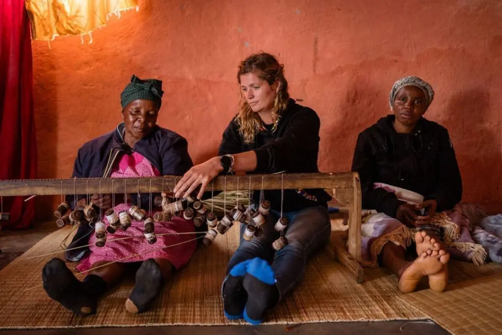 Drie vrouwen zitten binnenshuis op een mat tegen een oranje muur en werken samen aan een houten weefgetouw met meerdere spoelen draad. Twee vrouwen dragen hoofddoeken en traditionele kleding; één vrouw leidt het weven.