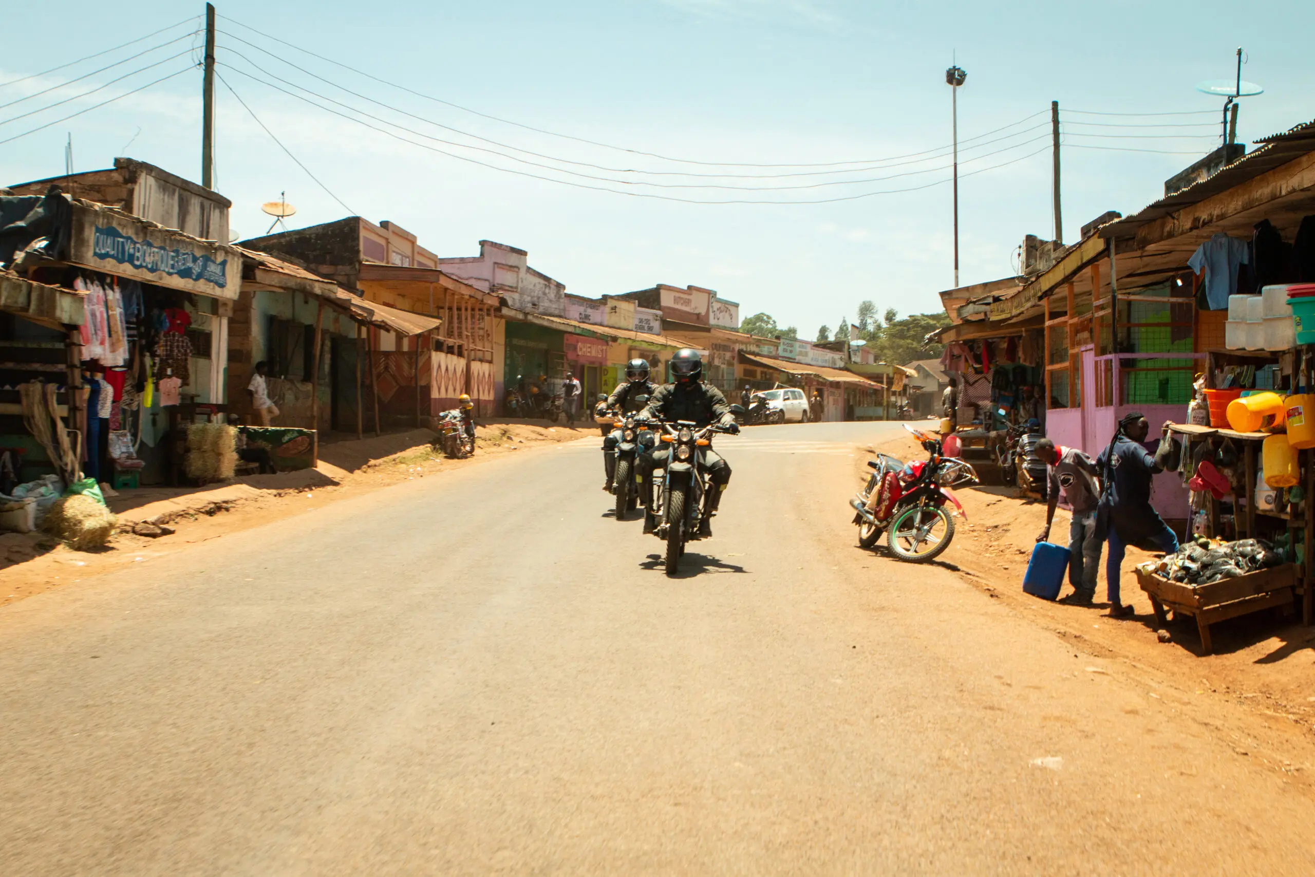 Drie motorrijders rijden door een zonovergoten, stoffige straat met marktkraampjes en kleine winkeltjes, terwijl voetgangers en geparkeerde motorfietsen langs de kant van de weg te zien zijn.