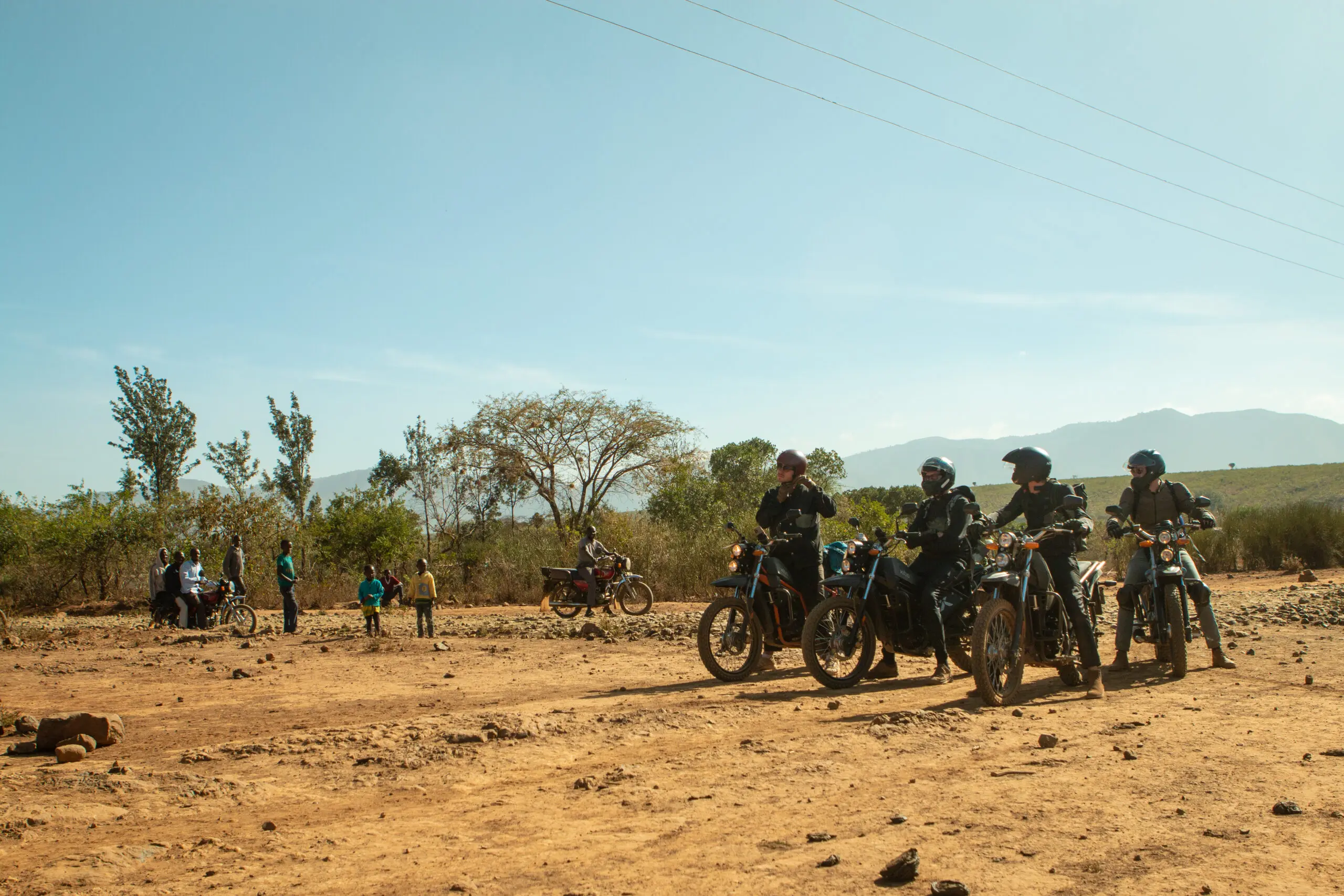 Vier mensen op motoren pauzeren op een droge, stoffige weg in een landelijke omgeving, terwijl een groep kinderen en volwassenen op de achtergrond staat onder een heldere, heldere hemel met verspreide bomen en heuvels in de verte.