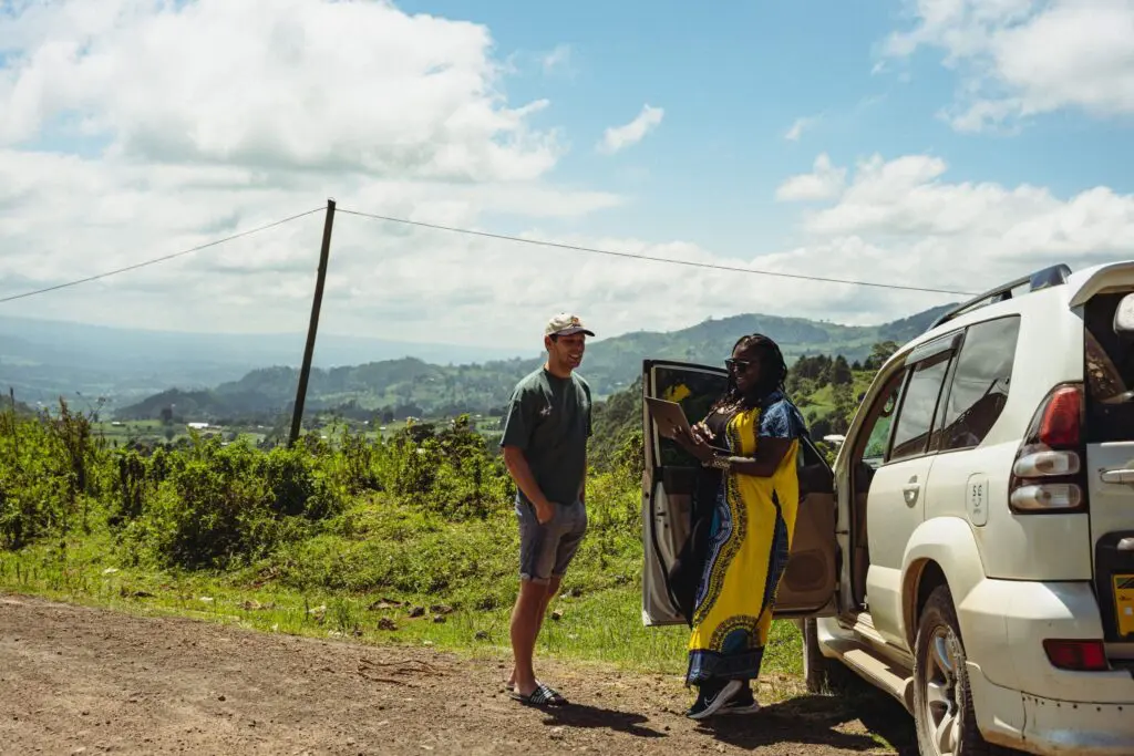 A man in casual clothing stands beside a woman in a vibrant yellow and blue dress, who is leaning on an open SUV door, with green hills and a blue sky in the background.