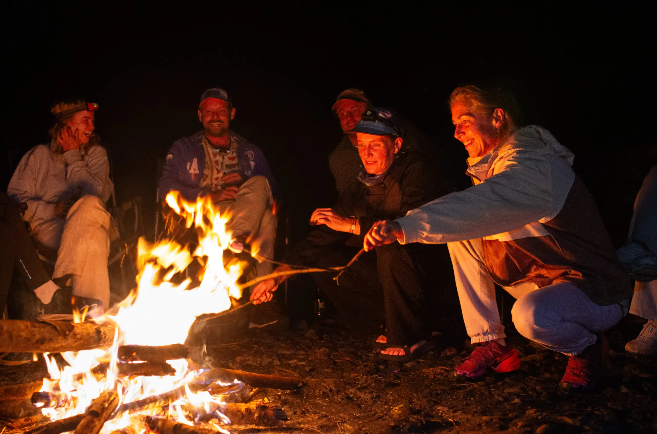 Een groep mensen zit en knielt 's nachts rond een kampvuur, glimlacht en roostert marshmallows boven de vlammen. Het warme vuur verlicht hun gezichten tegen de donkere achtergrond.
