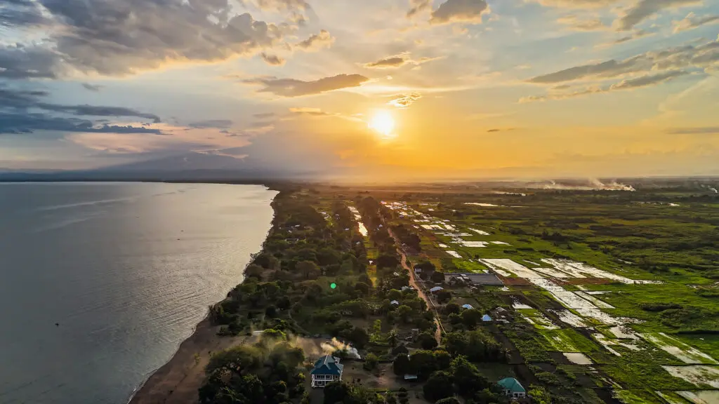 Luchtfoto van een kustlijn bij zonsondergang met de zon die door de wolken schijnt, water aan de linkerkant en groene velden en verspreide huizen aan de rechterkant. De lucht is kleurrijk met oranje, gele en blauwe tinten.