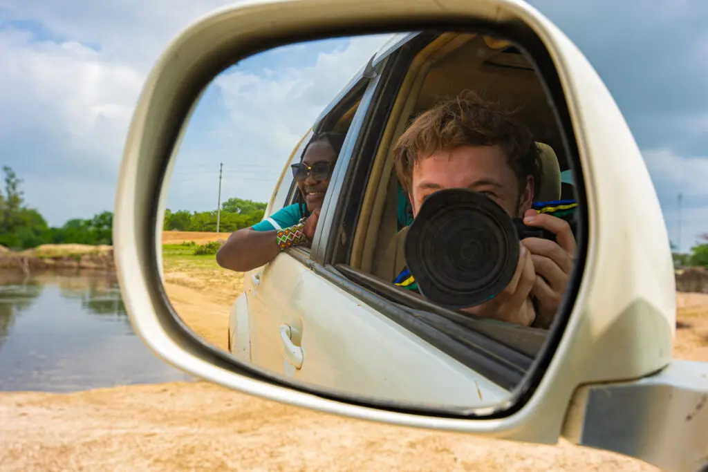 De zijspiegel van een witte auto weerspiegelt een man die een camera vasthoudt en een foto neemt, terwijl een andere persoon uit het autoraam leunt en glimlacht. De achtergrond toont water, zand en groen onder een bewolkte hemel.