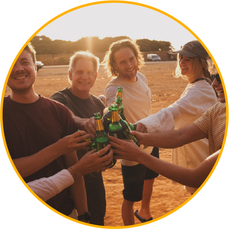 Een groep mensen die glimlachen en samen bierflesjes klinken bij zonsondergang, genietend van een informeel samenzijn of feest.