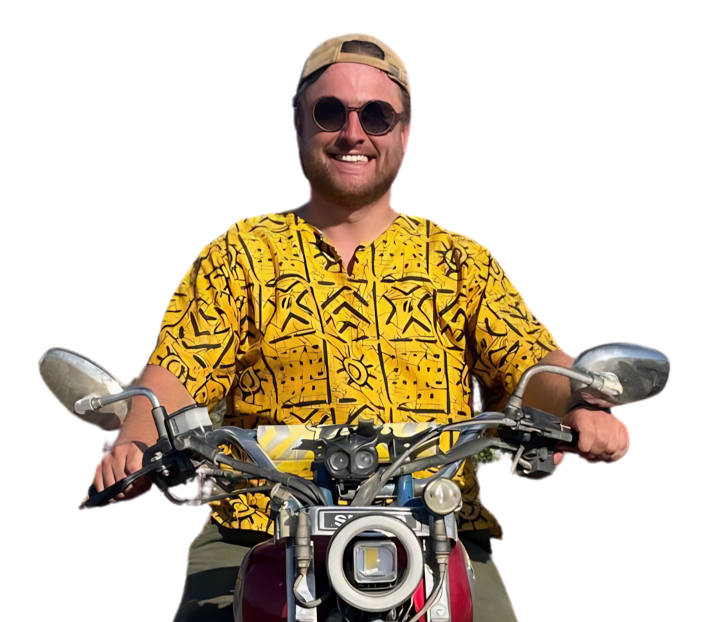 Een lachende man met zonnebril, beige pet en felgeel overhemd met patroon zit op een motorfiets, houdt het stuur vast, met een witte achtergrond.