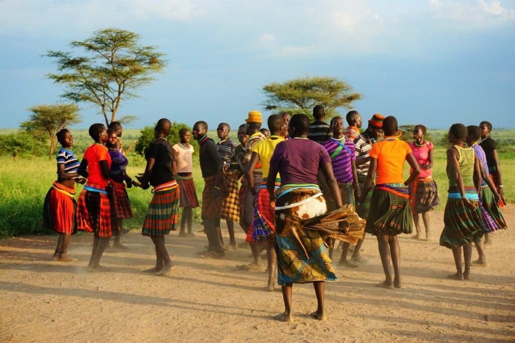 A group of people in colorful traditional clothing dance in a circle on a dusty path outdoors in Uganda, surrounded by green grass and trees under a bright African sky—an unforgettable scene for anyone travelling through Africa.