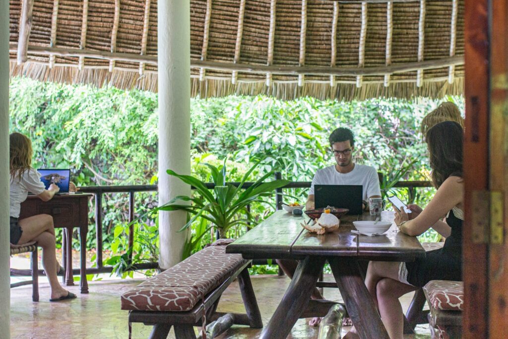 Four people work on laptops at wooden tables under a thatched roof, surrounded by lush African greenery. As they enjoy food bowls and drinks, their setup perfectly blends remote work with the spirit of travelling in Kenya.