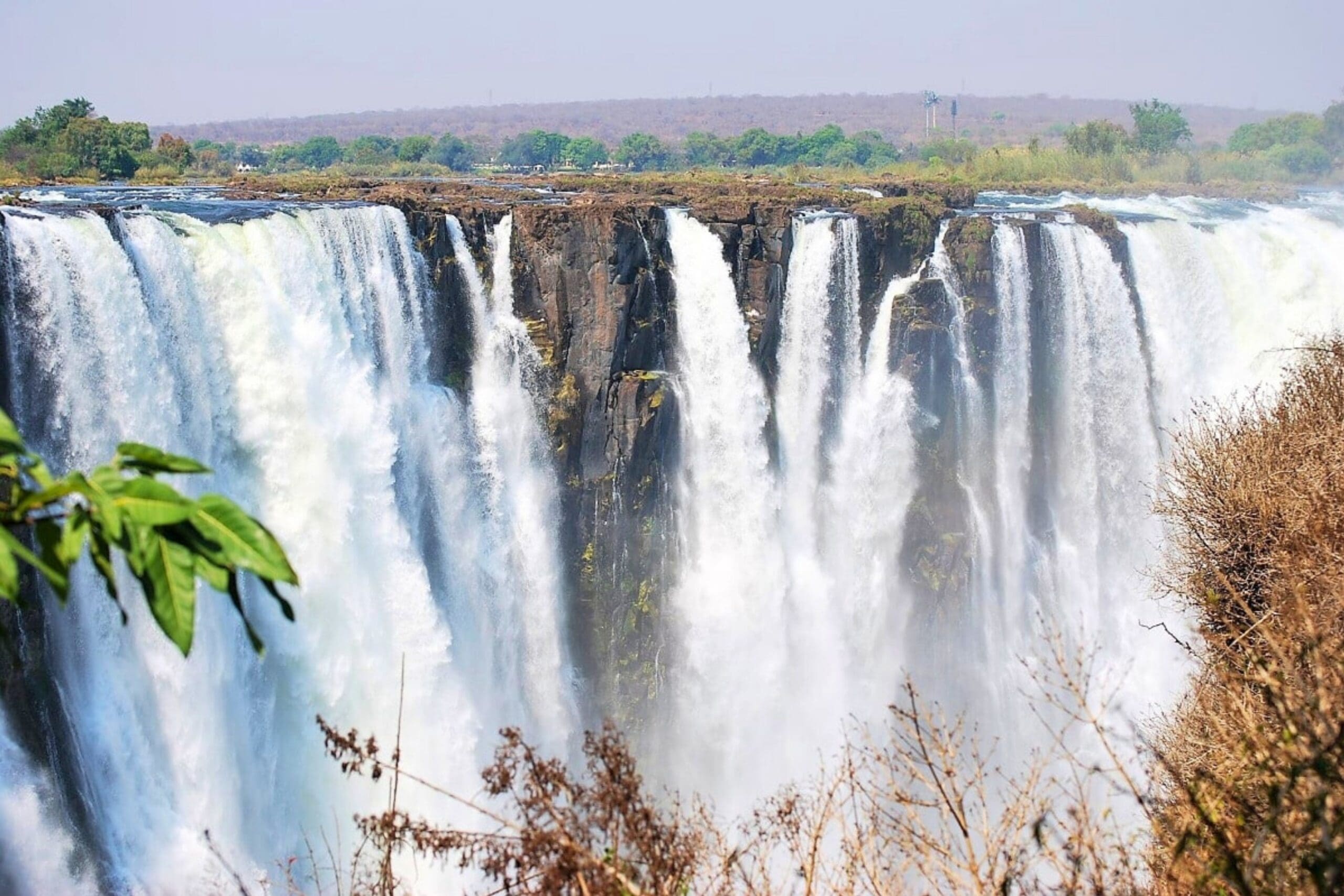 A wide view of powerful waterfalls cascading over a rocky cliff, surrounded by lush greenery and dry vegetation, with mist rising from below.