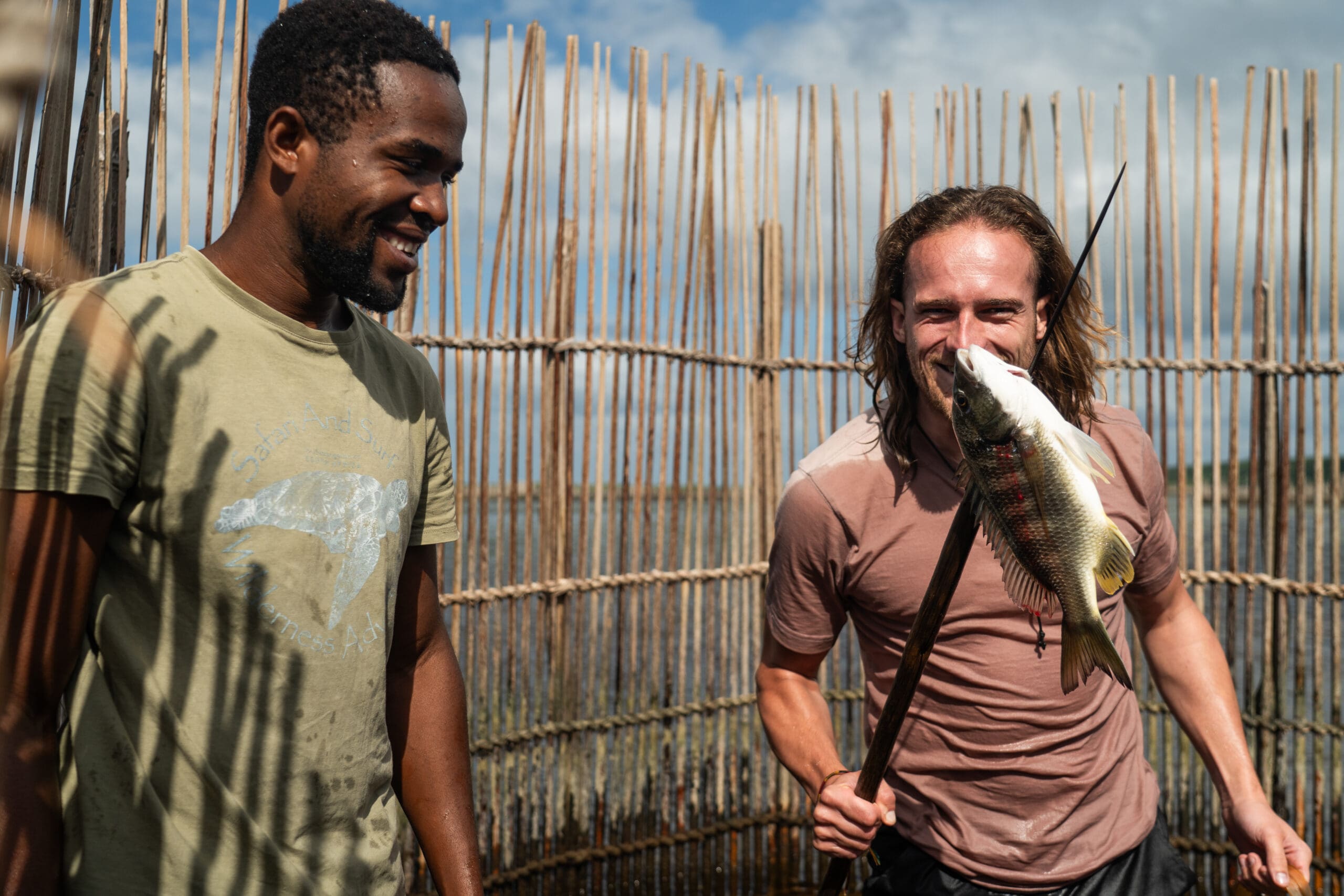 Twee mannen staan naast een bamboehek buiten in Zuid Afrika. De een glimlacht, terwijl de ander een speer met een vis erop vasthoudt. Het zonnige tafereel lijkt zich in de buurt van water of moerasgebieden te bevinden, wat de geest van het reizen in Afrika weergeeft.