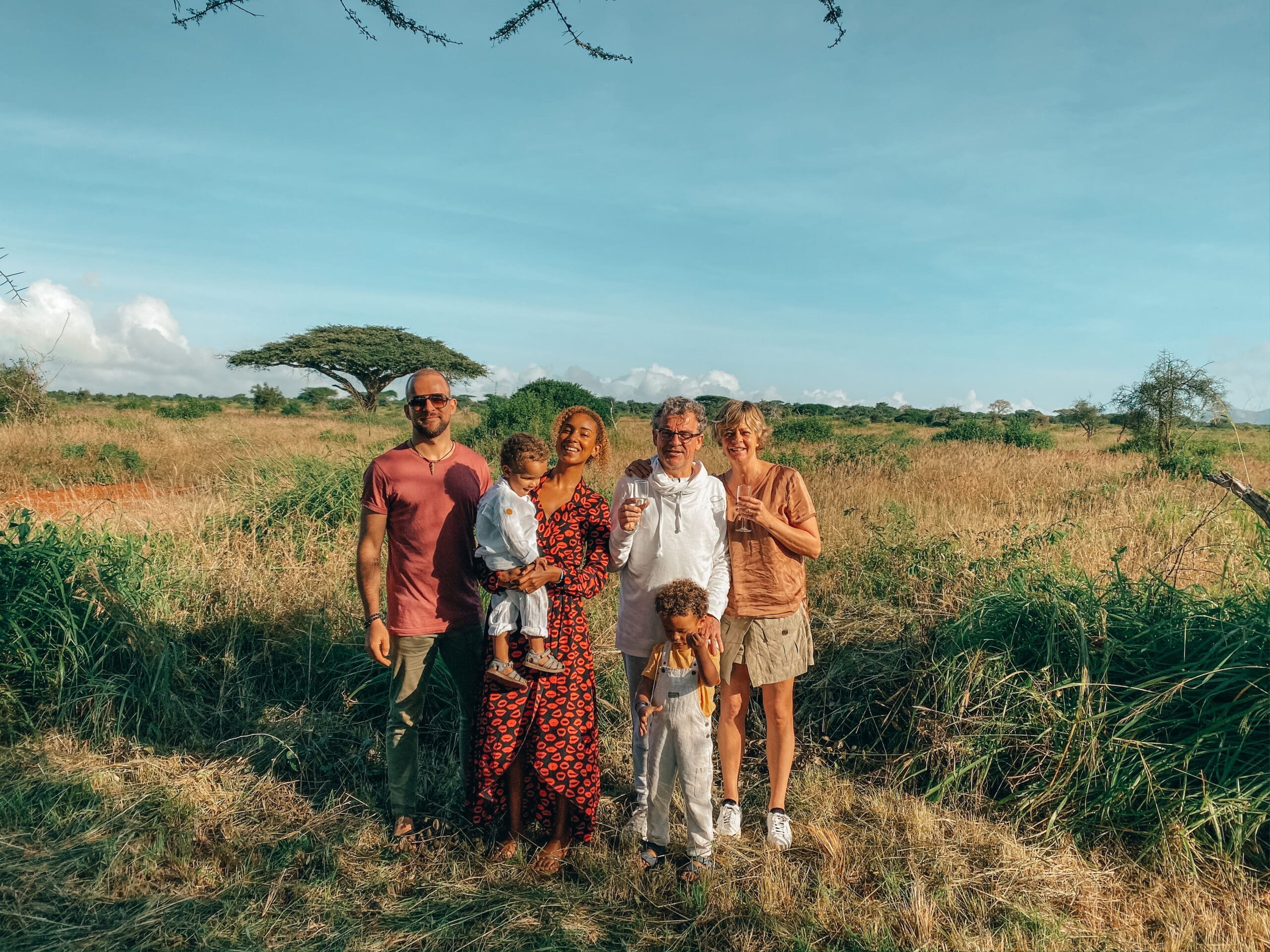 familie met jonge kinderen poseert in grasvlakte met wijnglazen in de hand tijdens safari in Kenia