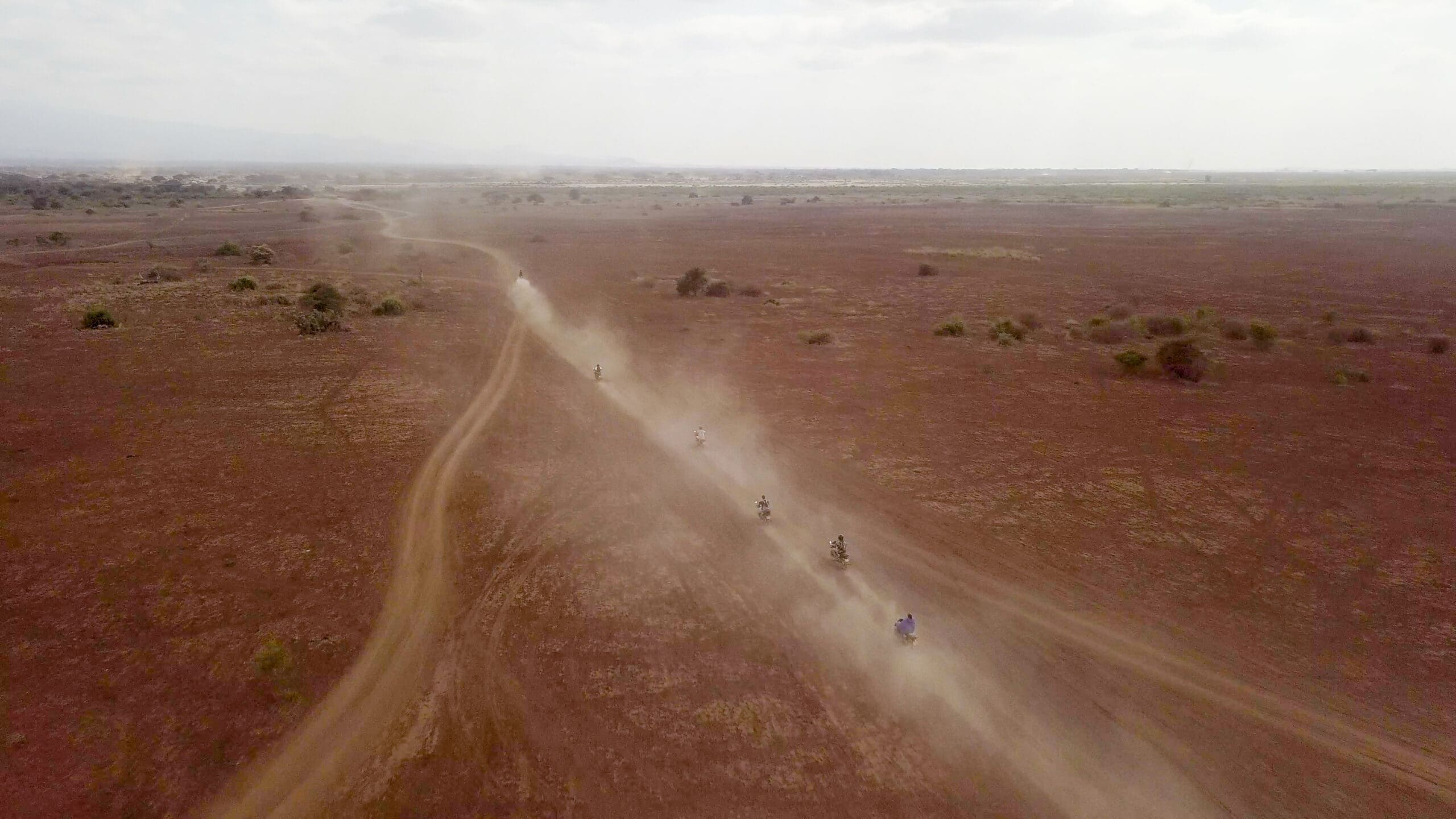 Luchtfoto van een droog, stoffig landschap in Tanzania met een kronkelige zandweg. Verschillende motorrijders rijden in een rij en laten stofsporen achter terwijl ze over het kale terrein rijden onder een bewolkte hemel.