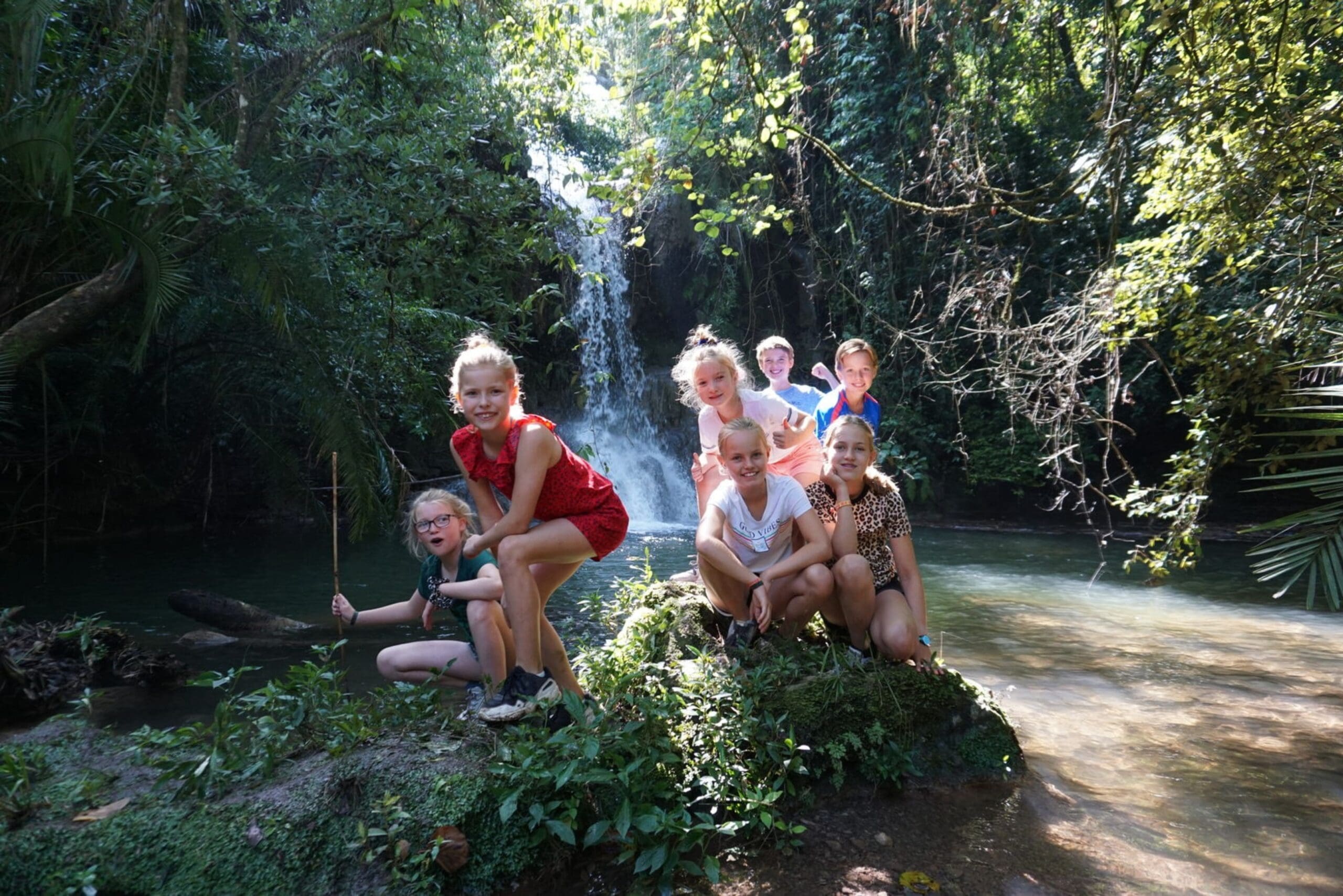 Zeven kinderen poseren op rotsen en groen voor een waterval en vijver in een weelderig, zonovergoten bos. Sommige kinderen hurken of zitten, terwijl anderen staan, allemaal glimlachend en genietend van de natuurlijke omgeving.