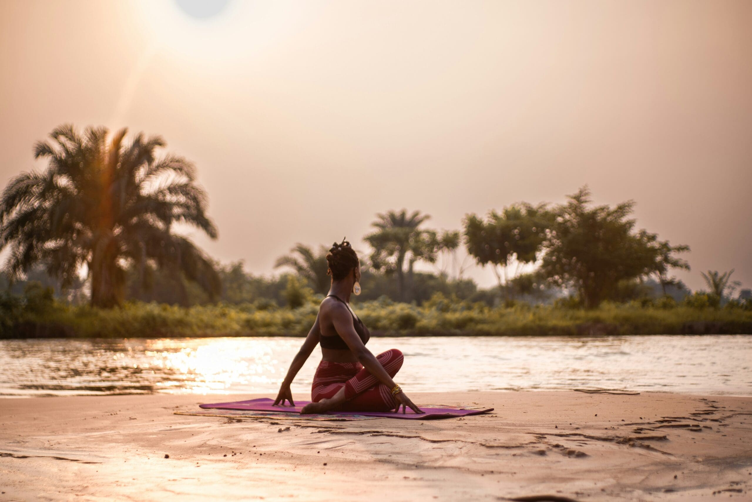 Een vrouw beoefent yoga op een matje bij een rivier tijdens zonsondergang, omringd door bomen en palmbomen onder een wazige hemel. Ze zit met haar rug naar de camera en kijkt naar het water.