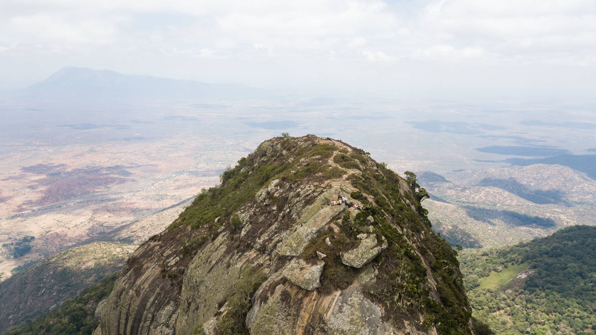 A rocky mountain ridge in Tanzania, covered with patches of green vegetation, rises sharply above a vast African landscape, with distant plains and hills visible under a partly cloudy sky—perfect for travelling adventurers.
