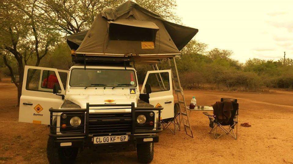 Een witte terreinwagen met een daktent staat geparkeerd op een onverharde ondergrond. Campingstoelen, een tafel met benodigdheden en bomen op de achtergrond suggereren dat Zuid Afrika reizigers genieten van een camping in een natuurlijke, droge omgeving.