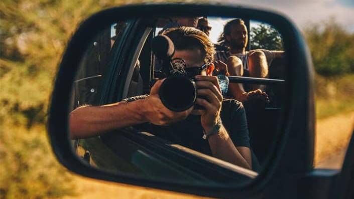 Een persoon met een camera wordt weerspiegeld in de zijspiegel van een auto en maakt een foto terwijl reizigers erin zitten. Op de achtergrond roepen wazig groen en zonlicht de geest van reizen op, wat doet denken aan een avontuurlijke reis in Afrika.