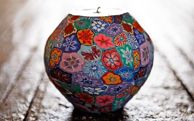 A round candle holder decorated with colorful, hand-painted floral patterns sits on a wooden surface, with light shining from behind it.