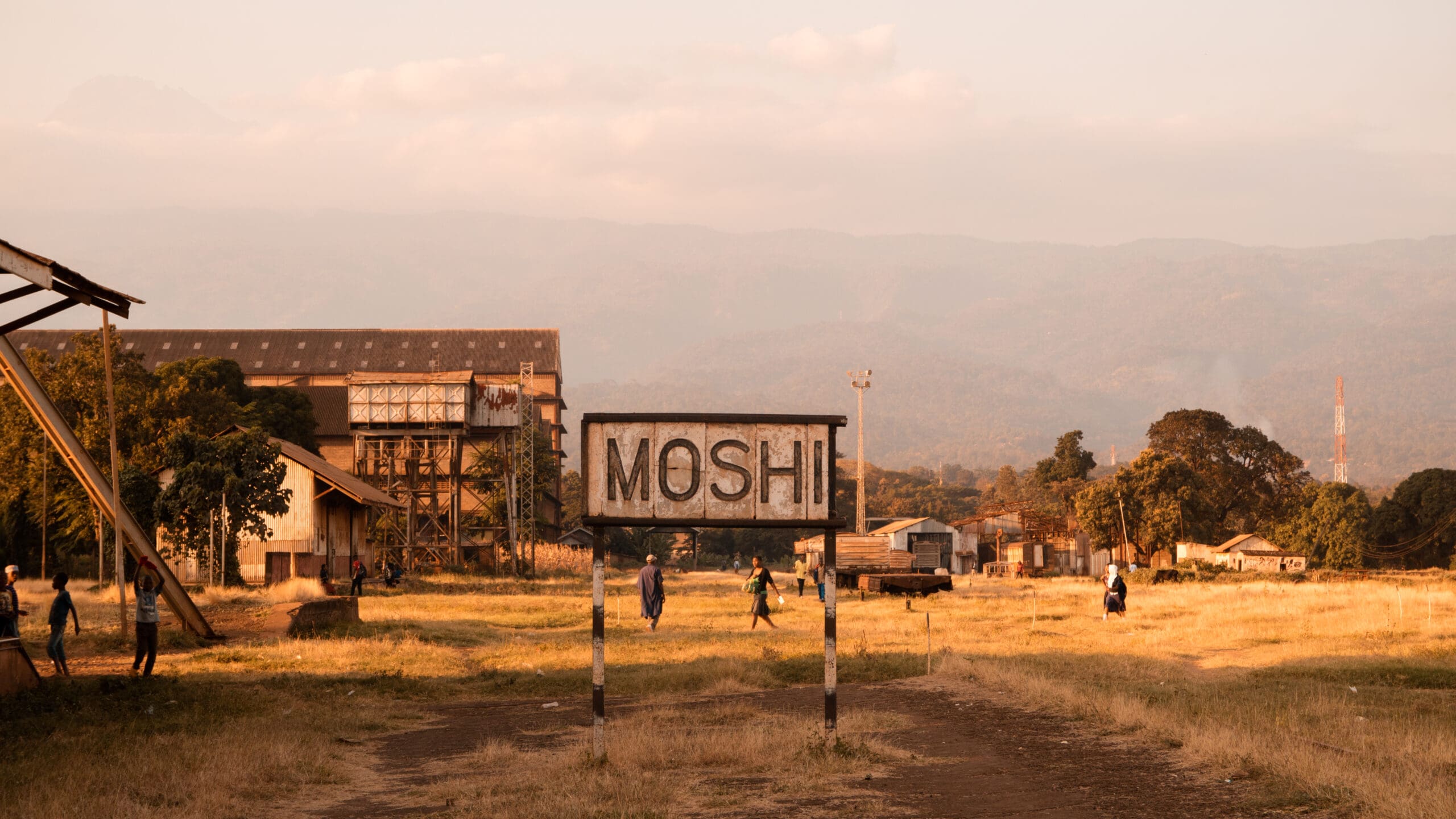 Een bord met de tekst MOSHI staat in een veld in Tanzania, met rondlopende mensen, oude industriële gebouwen, bomen en bergen in de verte onder een wazige Afrikaanse lucht.
