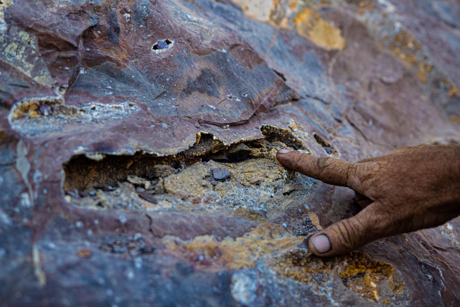 Een close-up van een persoonshand met een vuile vinger die wijst naar een ruw, veelkleurig rotsoppervlak met een diepe horizontale spleet en zichtbare minerale lagen.