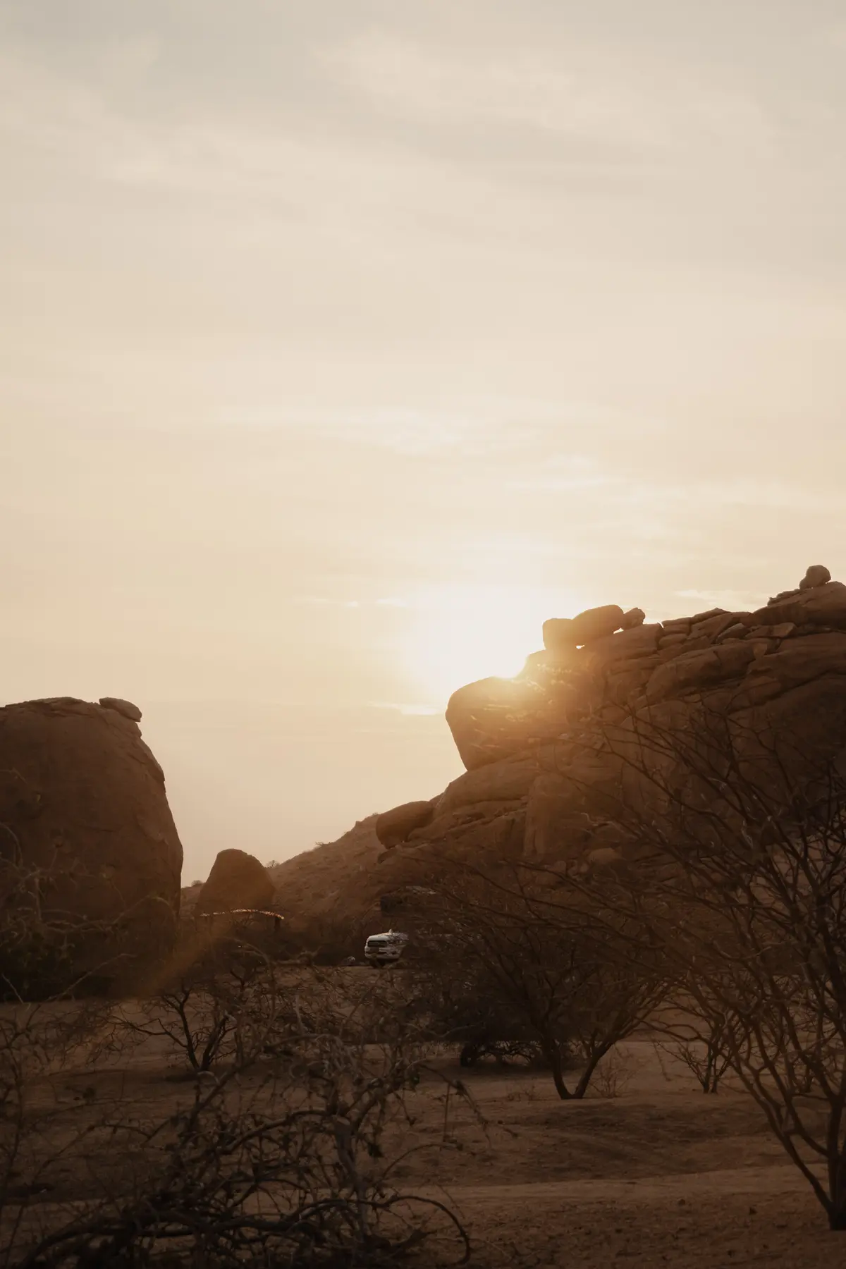 De zon gaat onder achter grote, rotsachtige heuvels en werpt een warm gouden licht over het landschap. Aan de voet van de rotsen op de voorgrond zijn enkele struiken en een eenzaam voertuig te zien.