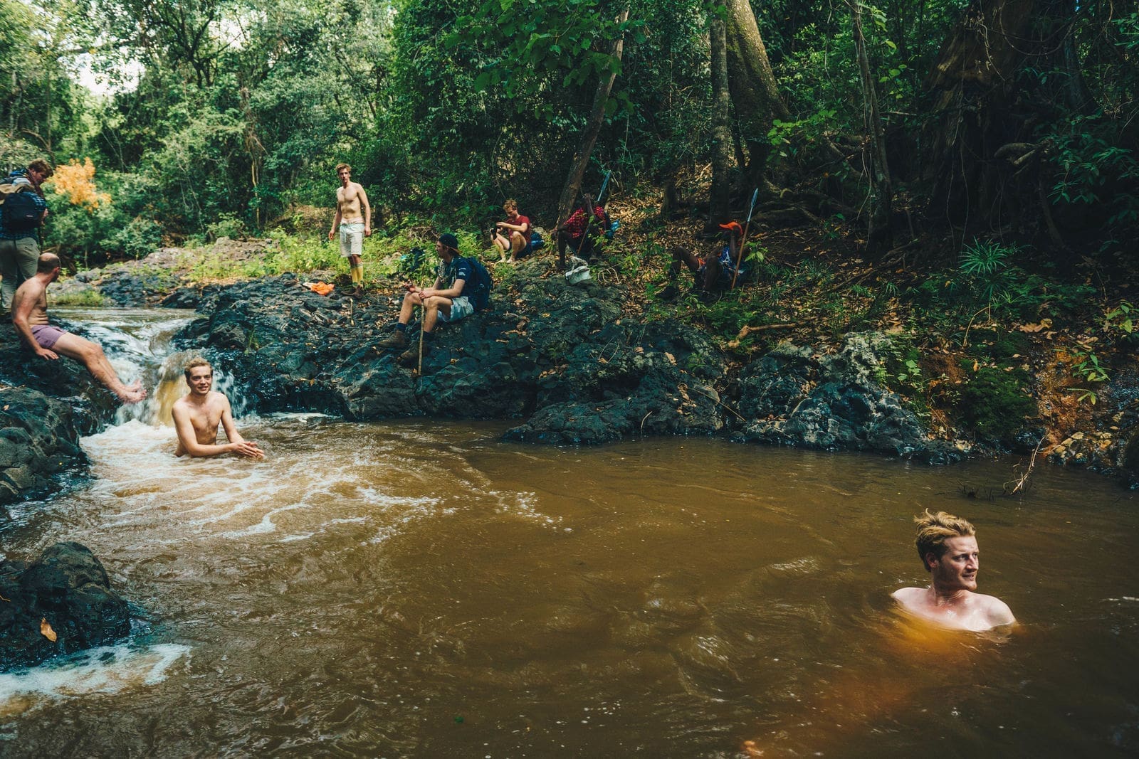 People relax and swim in a small, rocky stream surrounded by lush green trees in Kenya. Some sit on rocks while others wade or swim in the brownish water, enjoying the natural beauty of travelling through Africa’s scenic landscapes.
