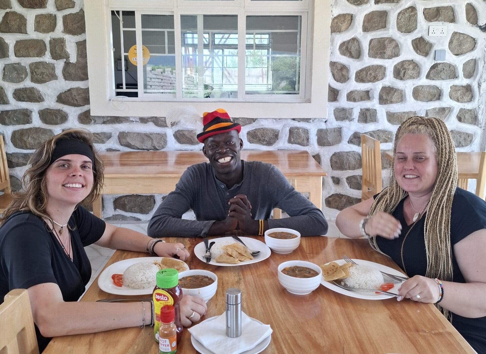 Drie reizigers zitten aan een houten tafel in Oeganda, glimlachen en genieten van een maaltijd van rijst en stoofpot in een lichte kamer met stenen muren en een raam op de achtergrond, die de warmte van Afrika vastlegt.