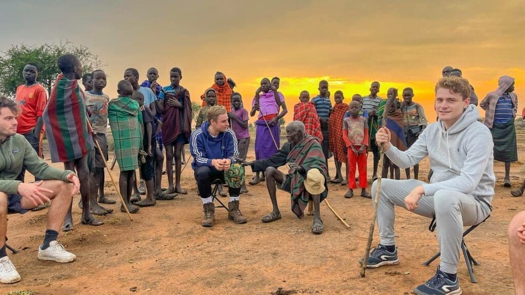 Bij zonsondergang in Oeganda verzamelen lokale stamleden in traditionele kleding en reizigers in vrijetijdskleding zich buiten, samen kletsend op de onverharde grond onder de uitgestrekte Afrika-hemel.