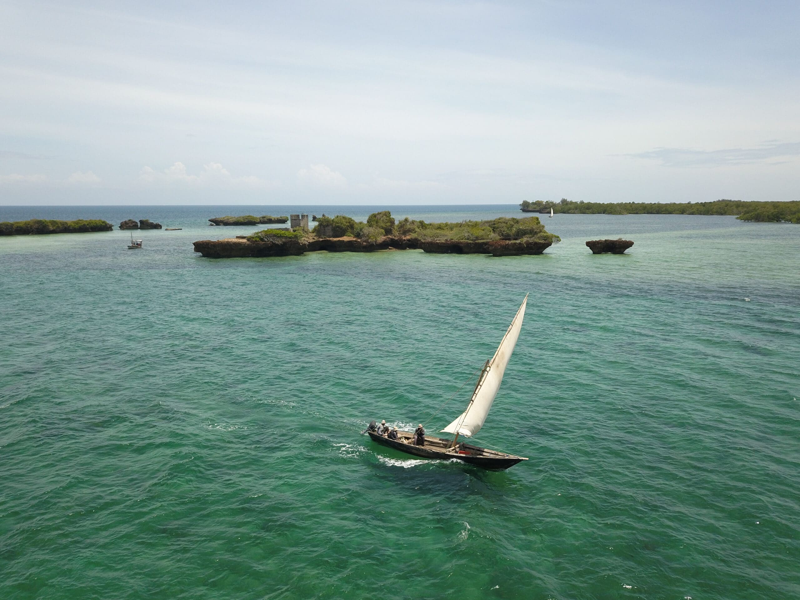 Ein kleines Segelboot mit weißem Segel gleitet über klares türkisfarbenes Wasser in der Nähe mehrerer felsiger, grüner Inseln unter blauem Himmel.