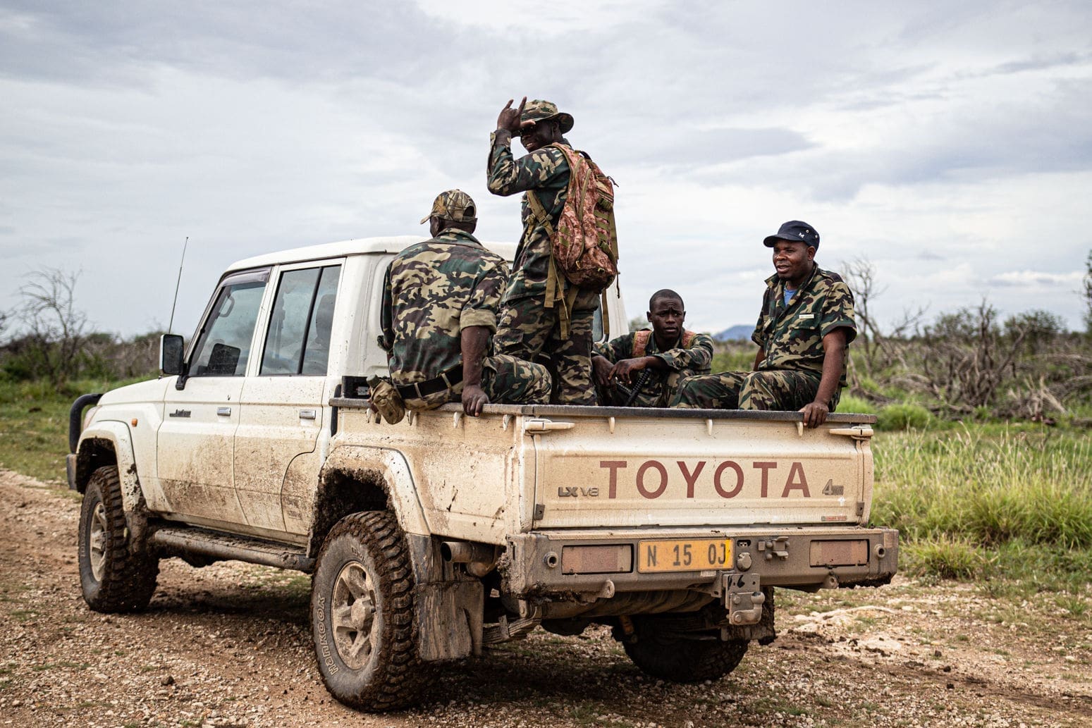 Four people in camouflage uniforms sit and ride in the back of a dusty white Toyota pickup truck on a dirt road in a rural area with grassy fields and cloudy skies.